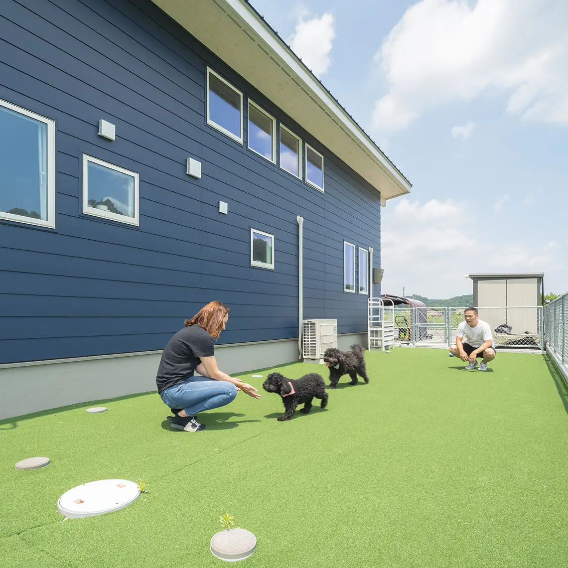 青い外壁の家の庭で、2人の大人が黒い犬と遊んでいる様子